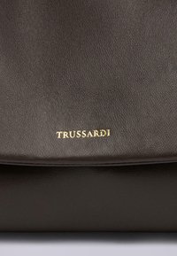 Tmavě hnědá kožená taška s hladkým povrchem a klopovým uzávěrem, na přední straně s vyraženým zlatým logem "TRUSSARDI".