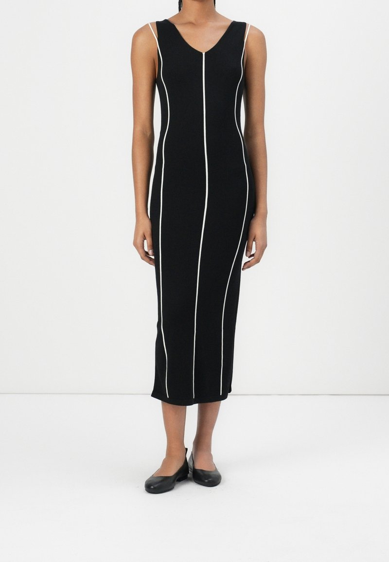 Robe midi noire sans manches avec un passepoil vertical blanc, portée avec des chaussures plates noires, debout devant un fond blanc uni.