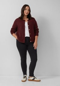 Bordový pletený kardigán s okrúhlym výstrihom a zapínaním na gombíky, nosený cez biele tričko, kombinovaný s čiernymi džínsami a viacfarebnými teniskami.