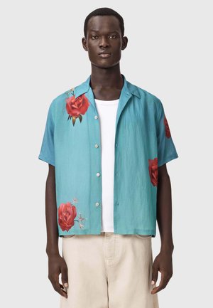 Ung mand iført en turkis kortærmet skjorte med røde rosenprint over en hvid t-shirt og beige bukser.