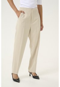 Pantalons beige ajustés avec poches latérales, design à jambes droite et subtils plis avant, associés à des escarpins noires à bout pointu.