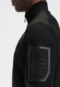 Juodas megztinis su užtrauktuku, aukštu apykakliu, pagamintas iš tekstūruotos mezgimo medžiagos, su lygiais odiniais akcentais ant pečių ir užtraukiama kišene ant rankovės.