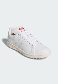 Vit lädersneaker med en slät yta, rund tå, snörningsdesign, perforerade sidodetaljer och en röd etikett på tungan.