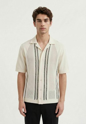 Ung mann iført kortermet kremfarget skjorte med svarte vertikale striper og mønstrede strikkepaneler, stående foran en ensfarget lys bakgrunn.