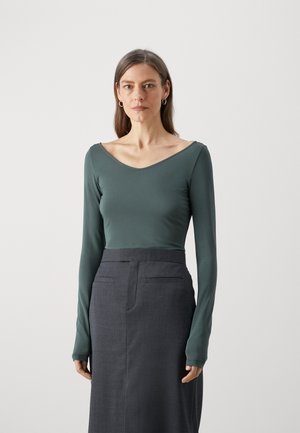 Top a maniche lunghe con scollatura a V profonda in tessuto verde tenue, abbinato a una gonna a tubino grigio scuro. L'outfit è aderente e di texture liscia.