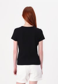 T-shirt nera a maniche corte con collo rotondo, mostrata da dietro. Tessuto liscio, design aderente, abbinata a pantaloni corti di jeans bianchi.