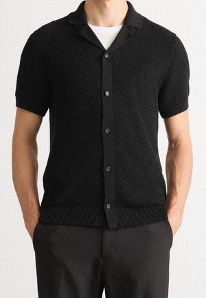 Hombre con cárdigan negro texturizado de manga corta abotonado sobre camiseta blanca y pantalones negros, de pie con las manos en los bolsillos.