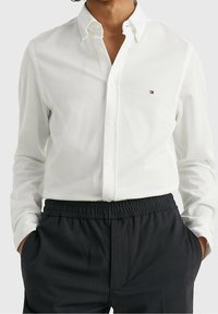 Camicia bianca a maniche lunghe in cotone, con colletto standard e un piccolo logo sul petto, abbinata a pantaloni scuri a fantasia.