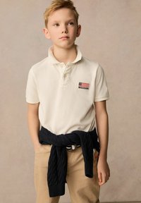 Krämfärgad polo med en liten amerikansk flagga på bröstet, ihop med beiga byxor. En marinblå stickad tröja är knuten runt midjan.