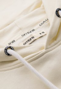 Gros plan sur un sweat à capuche couleur crème montrant un cordon blanc, des œillets métalliques noirs et des étiquettes de marque "ombre" à l'intérieur de l'encolure.