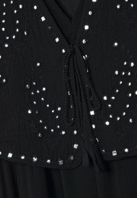Tissu noir avec broderie florale en perles argentées et sequins, et deux liens tressés avec pompons à l'encolure en V.