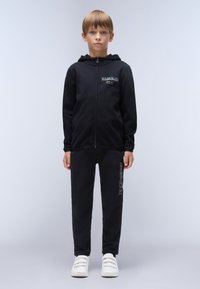 Zwarte zip-up hoodie met voorzak en logo detail, gecombineerd met zwarte sweatpants met tekst op de zijkant, gedragen met witte sneakers.