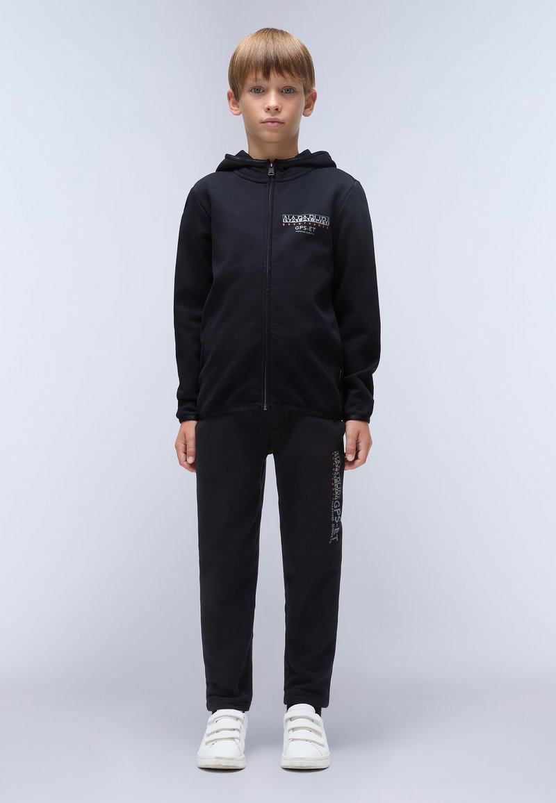 Zwarte zip-up hoodie met voorzak en logo detail, gecombineerd met zwarte sweatpants met tekst op de zijkant, gedragen met witte sneakers.
