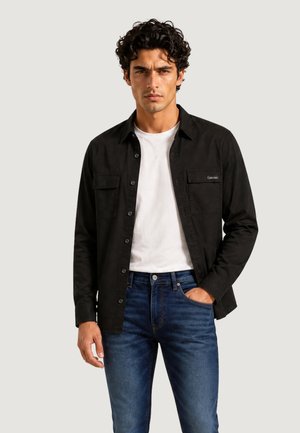 Man met krullend donker haar, een zwart hemd met knopen over een wit T-shirt en blauwe spijkerbroek, staat met één hand in zijn zak.