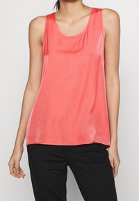 Korallfärgad ärmlös linne i ett mjukt, lätt tyg med rund halsringning och subtil lyster, kombinerad med svarta byxor.