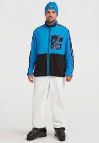 Chaqueta de cremallera azul y negra con patrones abstractos en rosa, combinada con pantalones blancos y botas de esquí negras. Acentuada con un gorro de lana azul.