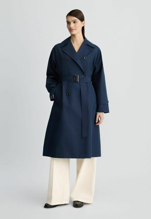 Navyblauwe trenchcoat met een ceintuur in de taille, dubbelgeknoopte voorkant, grote revers en wijde mouwen, gecombineerd met crèmekleurige wijde broekspijpen.