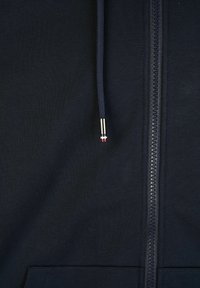 Dunkelblaues Zip-Hoodie aus glattem Stoff, mit einem durchgehenden Reißverschluss und einem silbernen Zipper mit rotem Akzent sowie einem Zugband am Kragen.