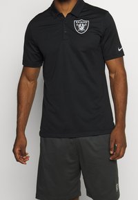 Mies, der ein schwarzes Raiders-Polohemd mit Logo auf der linken Brust und graue Sportshorts mit Logo auf dem linken Bein trägt, vor einem einfarbigen Hintergrund.