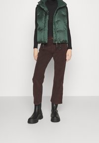 Chaleco acolchado verde con un cuello alto sobre una camiseta negra de manga larga, combinado con pantalones de pana burdeos y botas negras hasta el tobillo.