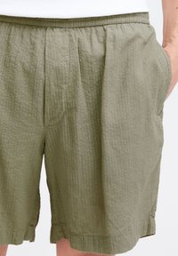 Pantaloni scurți verzi măsliniu, texturați, cu talie elastică și buzunare laterale. Prezintă dungi verticale și o croială lejeră.