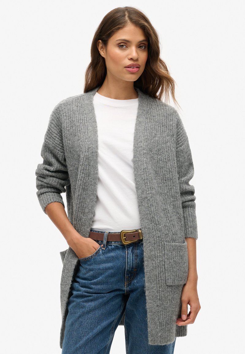 Cardigan en tricot gris avec une coupe décontractée, devant ouvert, manches longues et deux poches avant, associé à un t-shirt blanc et un jean bleu.