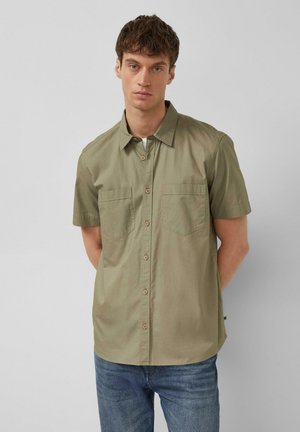 Jeune homme portant une chemise kaki à manches courtes avec deux poches poitrine et un jean bleu, debout devant un fond uni.