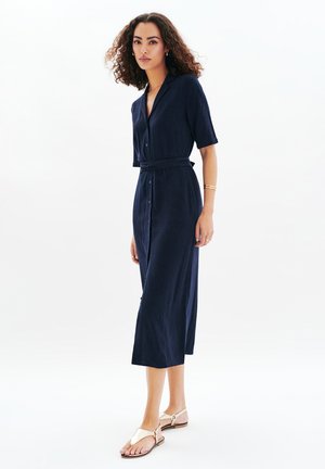Robe bleu marine à boutons, à manches courtes, avec une ceinture à la taille et une longueur midi. Associée à des sandales beiges, elle se distingue par un tissu lisse et texturé.