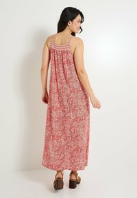 Robe maxi paisley rouge avec des fines bretelles, un dos froncé et un tissu fluide. Présente un motif et des talons épais bruns.