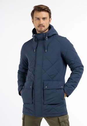 Mann trägt eine marineblaue, gesteppte Kapuzenjacke mit Klappentaschen vorne und Kordelzug an der Kapuze, steht mit den Händen in den Taschen.