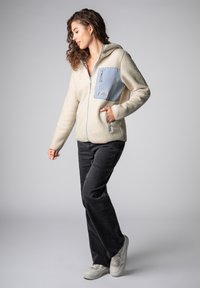 Vrouw in beige fleecevest met een blauwe borstzak, zwarte broek en witte sneakers, staand tegen een effen grijze achtergrond.