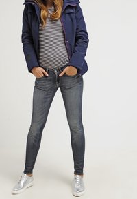 Giacca a vento imbottita blu scuro con cappuccio accentato in viola e una maglietta a maniche lunghe a righe. Jeans scuri slim-fit e sneakers argentate completano l'outfit.