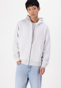 Felpa grigia chiara con zip, realizzata in tessuto morbido, con cappuccio, tasche frontali e polsini a coste. Abbinata a jeans blu chiaro.