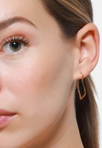 Orecchino a cerchio dorato con design a zigzag, caratterizzato da una finitura lucida. L'orecchino pende vicino all'orecchio di un modello, in armonia con uno sfondo neutro.