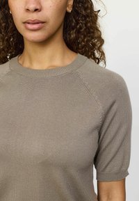 Bēšas krāsas īsām piedurknēm adīts tops ar apkakli un ribotām malām. Tam ir gluda tekstūra un raglanu piedurknes ar smalku šuvju detaļām.