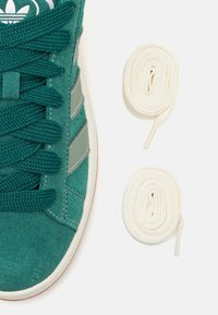 Zapatilla de ante verde con detalles de malla, suela de goma blanca y cordones planos blancos, dispuestos al lado del zapato en dos bobinas enrolladas.