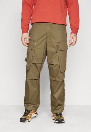 Pantalones cargo en verde oliva, hechos de tejido duradero; cuentan con múltiples bolsillos, detalles de cremallera y un corte holgado. Llevados con zapatillas negras y naranjas.