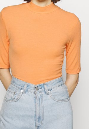 Person trägt ein oranges kurzärmeliges Shirt mit Stehkragen, das in hoch taillierte hellblaue Jeans mit Vordertaschen gesteckt ist.