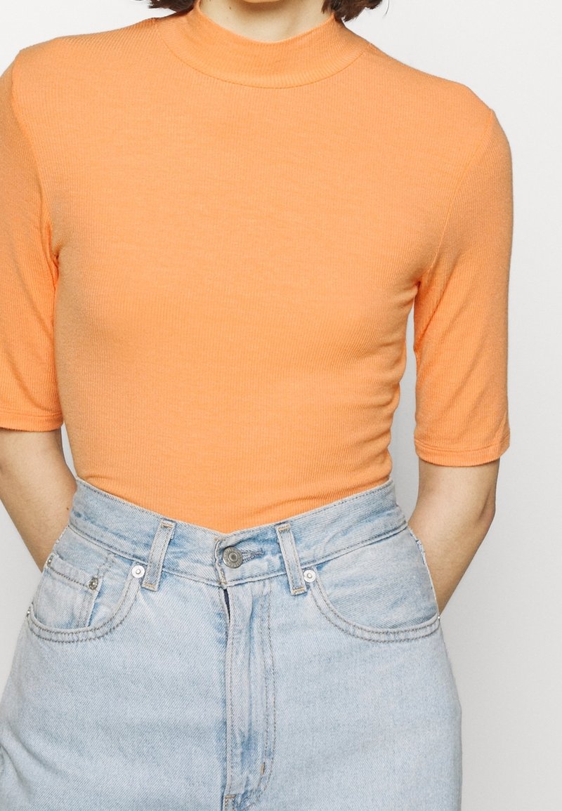 Personne portant un haut orange à manches courtes avec col montant, rentré dans un jean taille haute bleu clair avec des poches devant.