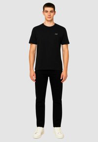 T-shirt in cotone nero con scollo rotondo, caratterizzato da un piccolo logo sul lato sinistro del petto, abbinato a jeans neri e sneaker bianche.