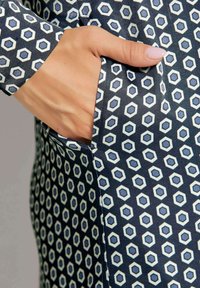 Marineblaues Gewebe mit einem hexagonalen Muster in Weiß und Hellblau; verfügt über eine Seitentasche und eine glatte Textur. Mit der Hand in die Tasche gelegt.