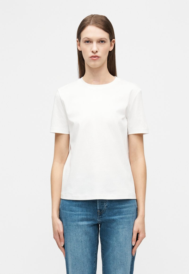 Γυναίκα με ίσια καστανά μαλλιά, που φοράει απλό λευκό κοντομάνικο μπλουζάκι και μπλε τζιν τζιν, στέκεται μπροστά σε λευκό φόντο.