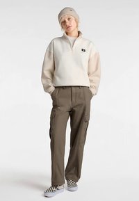 Sweat-shirt à fermeture éclair crème et beige, pantalon cargo olive, et chaussures à enfiler à carreaux. Le modèle porte un bonnet beige clair et pose sur un fond neutre.