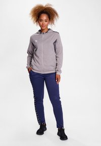 Grijze rits hoodie met een capuchon en donkere accenten, gecombineerd met marineblauwe trainingsbroek met een zigzagpatroon aan de zijkant, gedragen met zwarte sportschoenen.