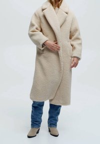 Manteau en laine beige texturée avec un large col, des poches avant et une coupe ouverte, porté sur un jean bleu et des bottines beige.