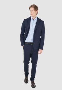 Abito blu navy con giacca aderente e pantaloni abbinati. Camicia azzurro chiaro sotto, abbinata a una cintura nera strutturata e scarpe marroni.