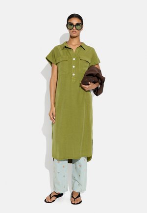 Kobieta nosi oversize zieloną sukienkę koszulową, jasnoniebieskie kwiatowe spodnie, czarne sandały, duże okrągłe okulary przeciwsłoneczne, trzyma złożoną brązową kurtkę.