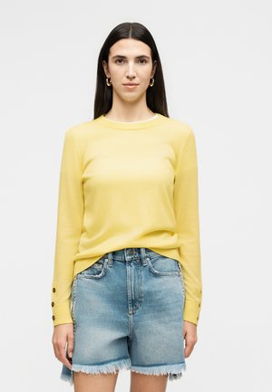 Naine, kellel on pikad tume juuksed, kuldsed rõnga-kujulised kõrvarõngad, kollane pika varrukaga kampsun ja kulunud äärega teksapüksid, ühevärvilise taustaga.