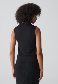 Zwarte mouwloze top van een textuurstof met een gekartelde halslijn. Het ontwerp is nauwsluitend en reikt tot de taille, waardoor een eenvoudige stijl wordt benadrukt.