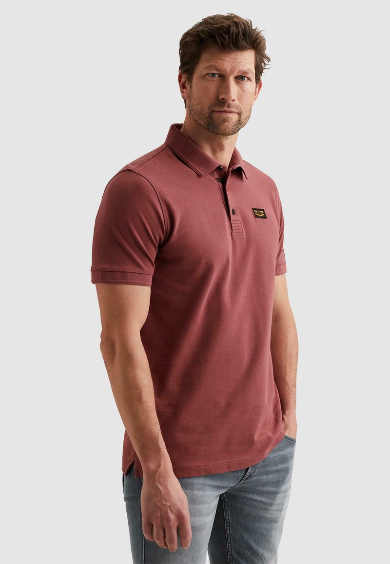 Homme portant un polo rouge brique à manches courtes avec un petit patch logo noir et or sur la poitrine, associé à un jean bleu clair.